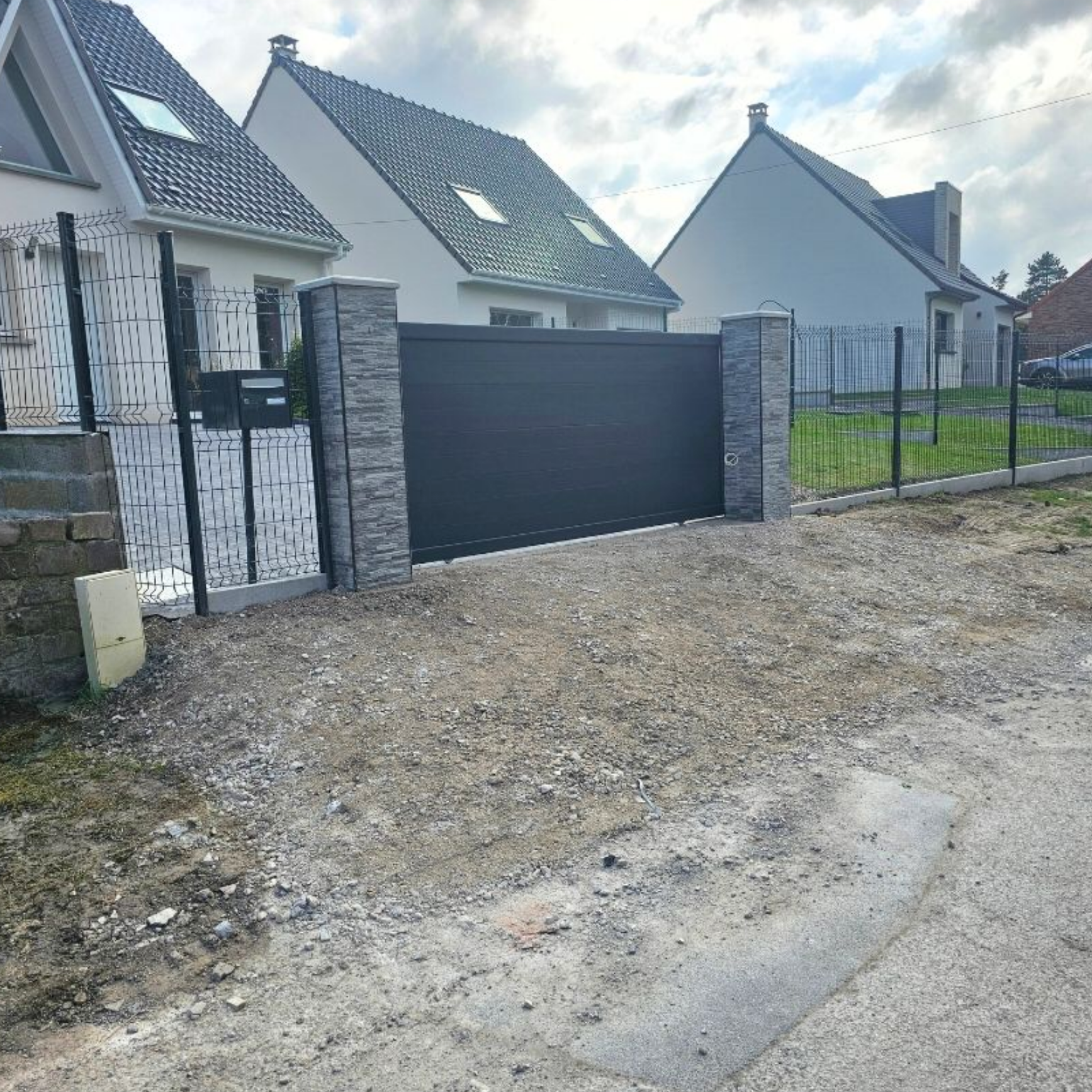 Portail noir devant des maisons avec allée de gravier, piliers en pierre et clôture grise. Ciel couvert.