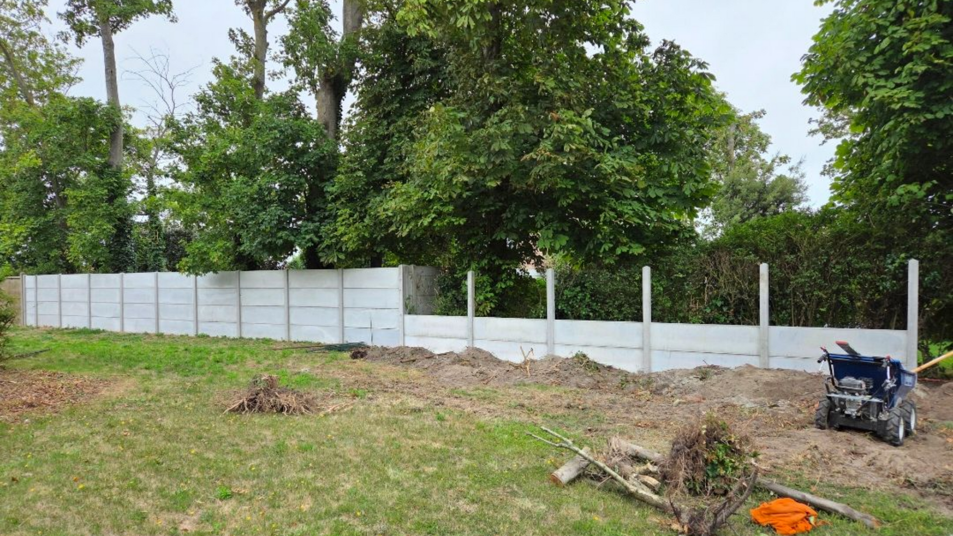 Construction d'une clôture en béton dans une cour herbeuse, avec des arbres en arrière-plan et une chargeuse compacte.