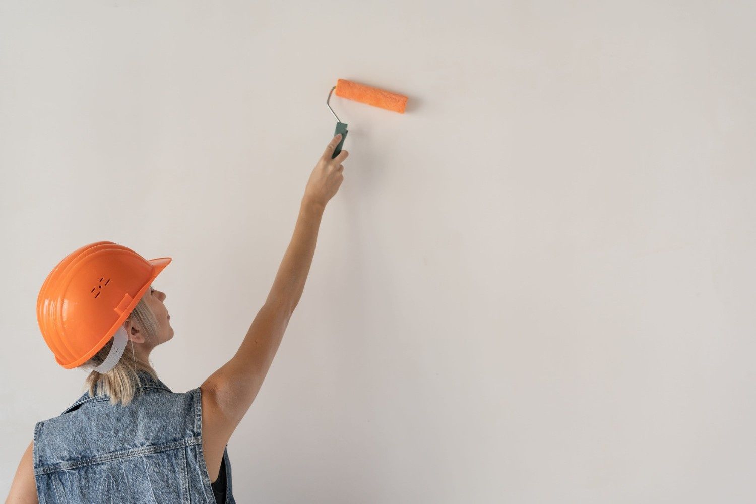 Una persona que lleva un casco naranja y un chaleco vaquero utiliza un rodillo de pintura para pintar una pared blanca lisa.