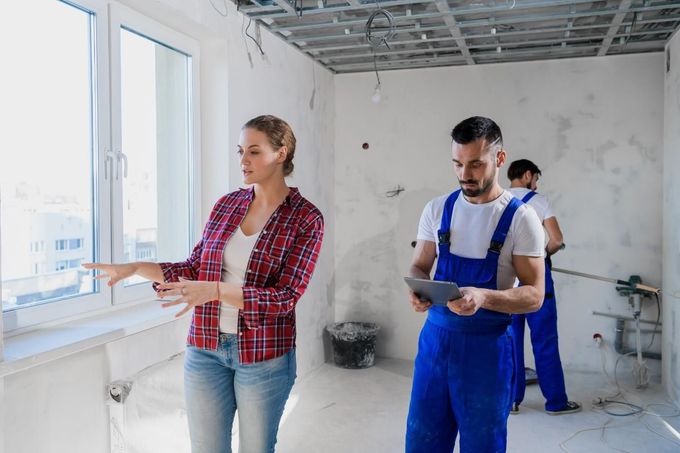 Una persona señala una ventana mientras un trabajador con mono azul consulta una tableta en una habitación en obras.