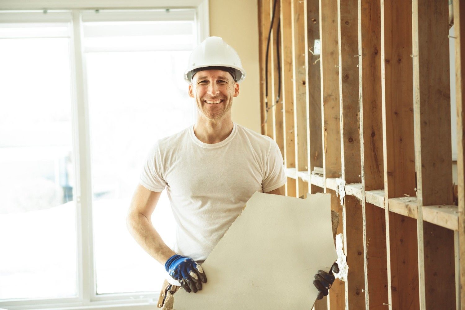 Un trabajador sonriente, con casco blanco y camiseta, sostiene un trozo de placa de yeso en una habitación en construcción.