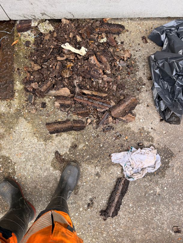 Tas de compost avec des bâtons et des débris sur du béton, à côté d’un sac-poubelle, vu de dessus avec des bottes visibles.