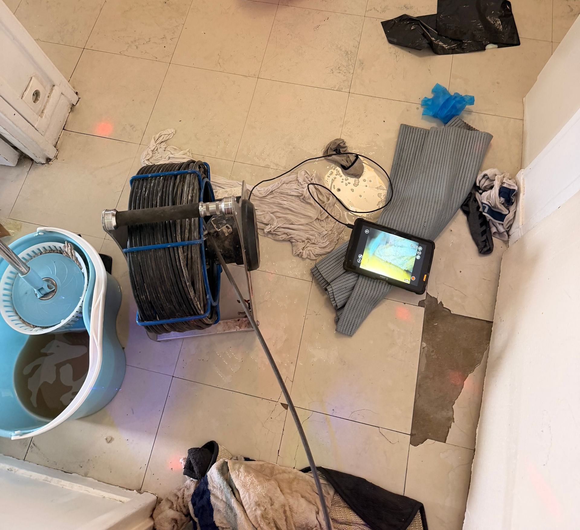 Pièce carrelée avec un enrouleur de câble, un seau de lavage et une tablette posés au sol parmi des outils et des débris éparpillés.