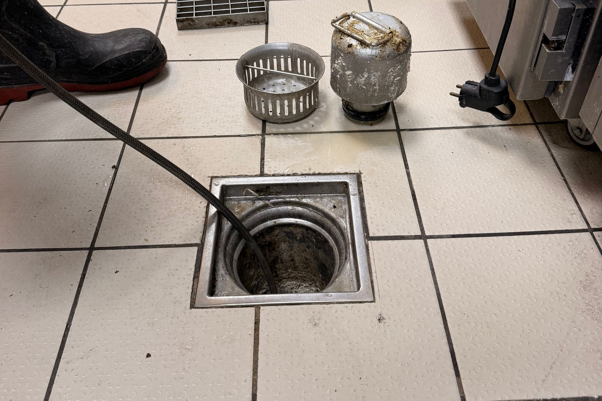 Caniveau de sol ouvert dans le carrelage d’une cuisine, à côté de filtres métalliques et de la botte d’un employé.