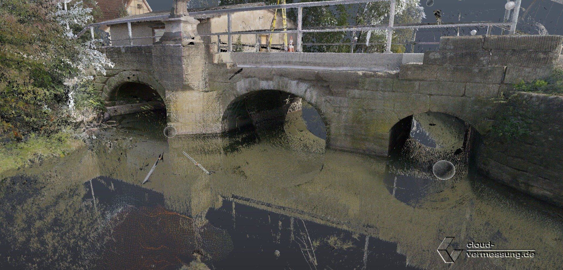 Steinbrücke über Wasser, mit Bögen und Gebäuden im Hintergrund. Reflektierende Wasseroberfläche.