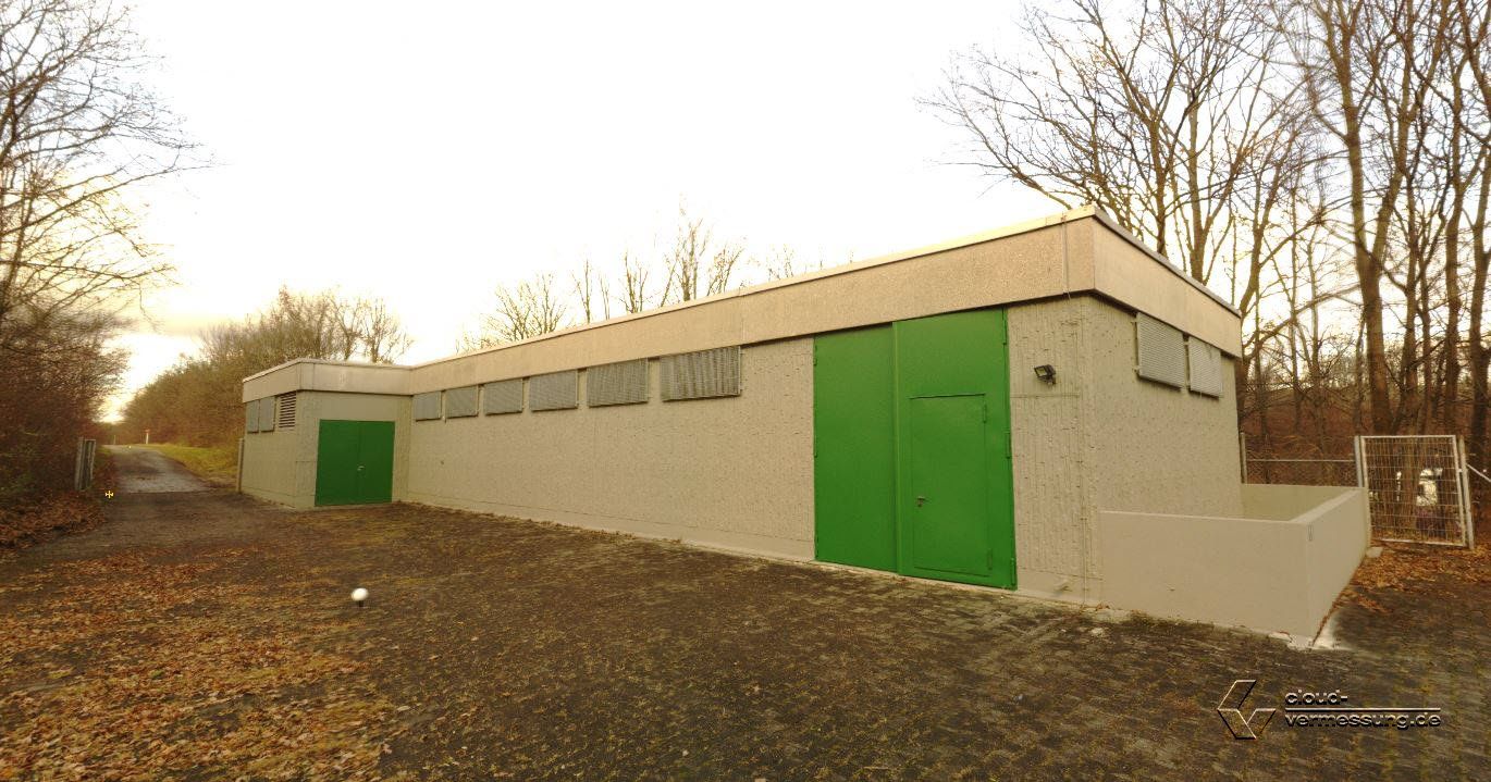 Außenansicht eines hellen Industriegebäudes mit grünen Türen und Fenstern, Kiesweg und Bäumen.