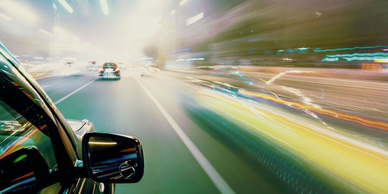 Auto fährt nachts, verschwommene Lichter ziehen vorbei. Fokus auf Seitenspiegel und Straße vor uns.
