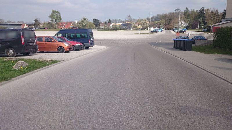 Asphaltierte Straße mit geparkten Autos auf der linken Seite und einem Parkplatz weiter vorne.
