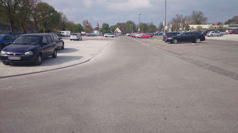 Parkplatz mit geparkten Autos, einer Straße und Gebäuden im Hintergrund unter einem bewölkten Himmel.