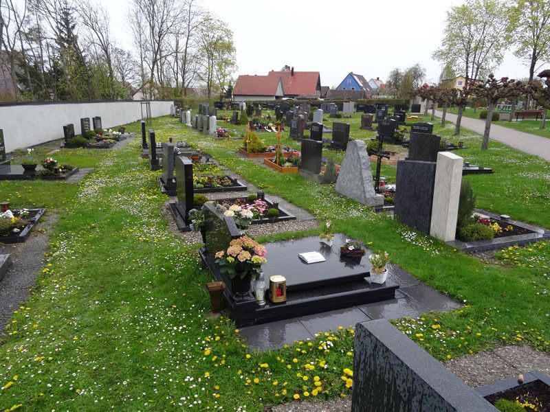 Friedhof mit Grabsteinen und blühenden Blumen, grünem Gras und Wohngebäuden im Hintergrund.