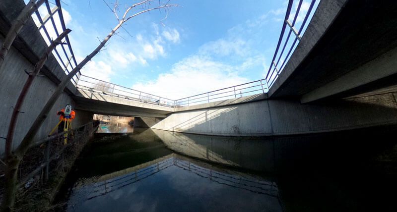 Untersicht einer Betonbrücke über einen ruhigen Fluss an einem sonnigen Tag; links ist eine Person zu sehen.