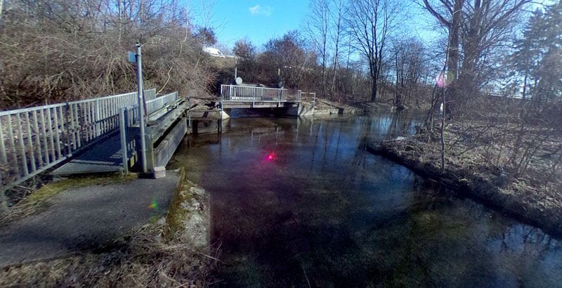 Ein Bach fließt unter einer Holzbrücke hindurch, im Hintergrund sind Bäume und ein Gebäude zu sehen. Sonniger Tag.