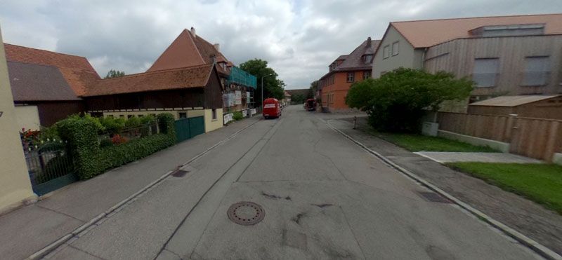 Straße in einem Dorf mit Häusern auf beiden Seiten und einem roten Fahrzeug in der Ferne unter einem bewölkten Himmel.