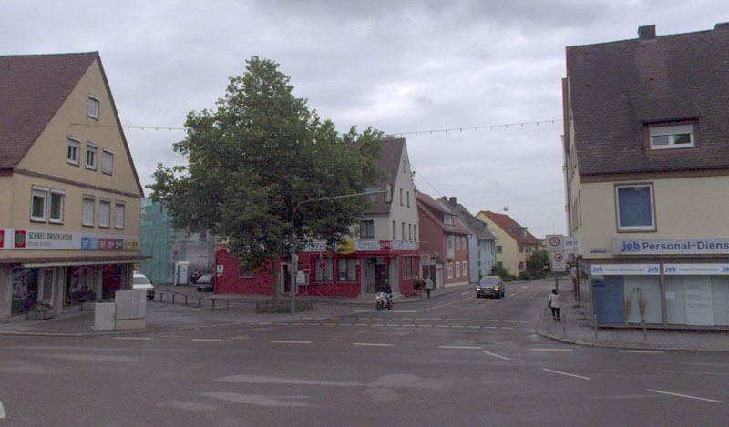 Straßenecke mit Gebäuden und einem Baum unter einem bewölkten Himmel. Ein Auto fährt durch die Kreuzung.