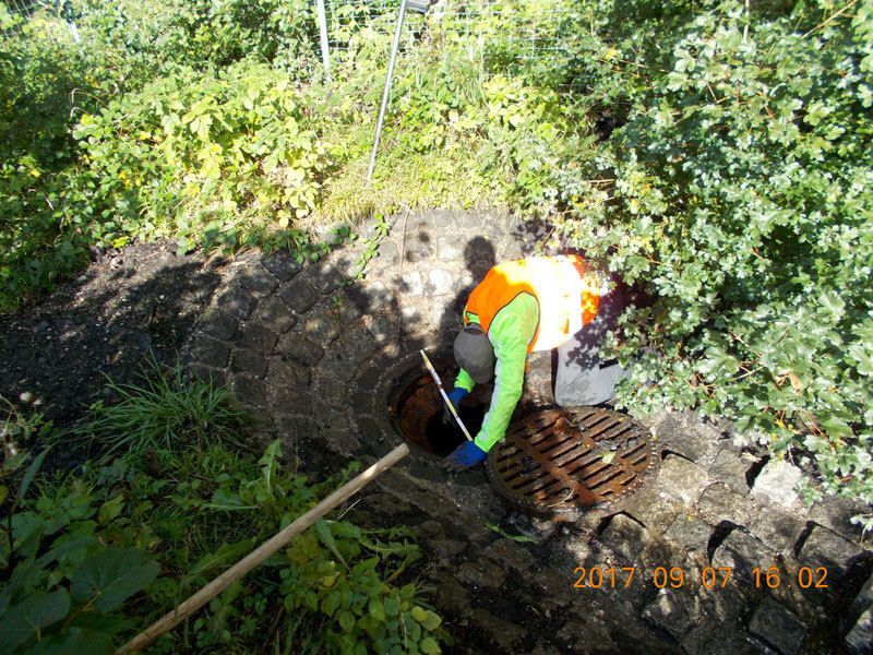 Eine Person in einer orangefarbenen Weste reinigt ein Abflussgitter in einem grasbewachsenen Außenbereich.