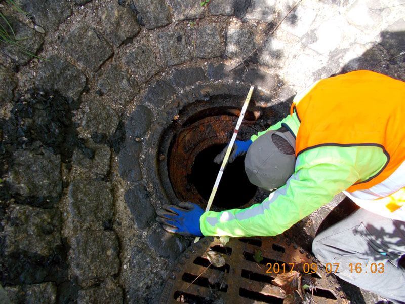 Person in Warnweste inspiziert offenen Schacht mit Messstab.