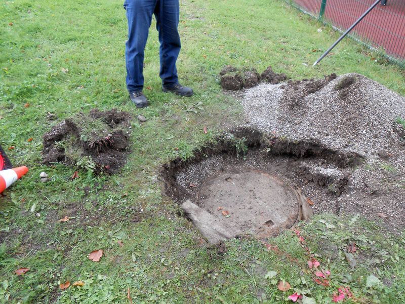 Person steht neben einer ausgegrabenen kreisförmigen Betonstruktur in einer Rasenfläche; um das Loch herum sind Kies- und Erdhaufen.