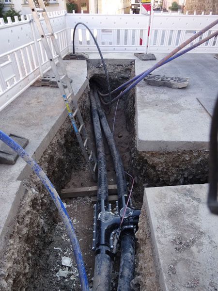 Baustelle: Kabel im Graben, schwarz und blau, mit elektrischem Anschlusskasten. Weißer Zaun und Leiter vorhanden.