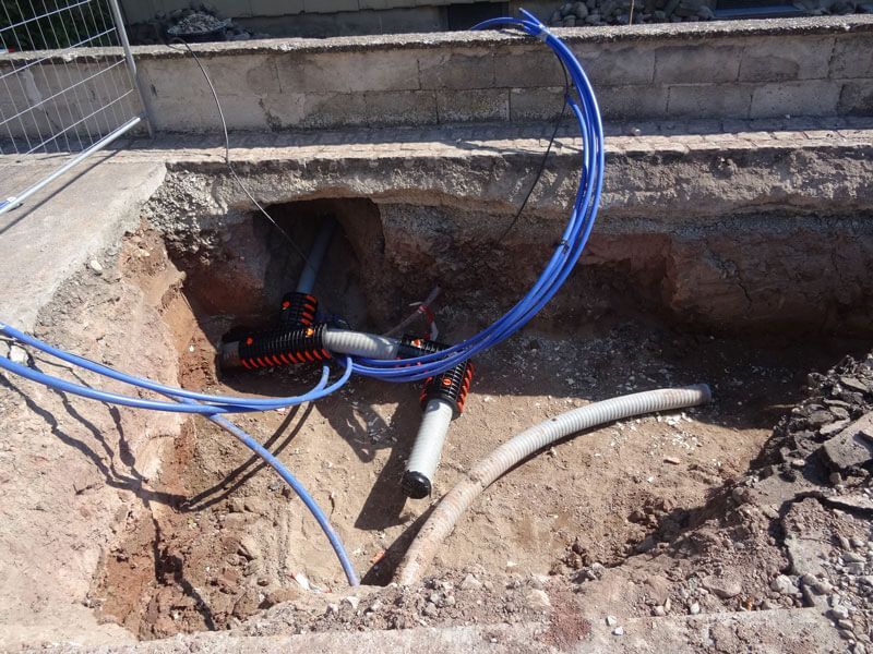 Ausgegrabener Boden zeigt blaue Rohre, elektrische Leitungen und Anschlüsse.