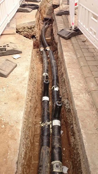 Schwarze Kabel in einem Graben, möglicherweise für Versorgungseinrichtungen, neben einem gepflasterten Bereich und Bauabsperrungen.