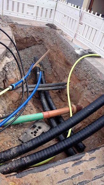 Offener Graben mit verschiedenen unterirdischen Versorgungsleitungen; schwarz, blau, grün und gelb.