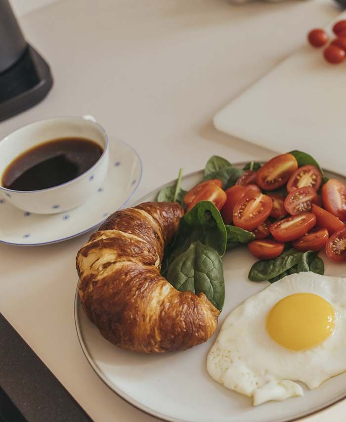 Un plato de comida con un croissant, huevos, tomates y espinacas junto a una taza de café.