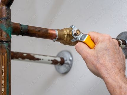 Eine Hand dreht ein Ventil mit gelbem Griff an einem Kupferrohr, das wahrscheinlich den Wasserfluss regelt und vor einer weißen Wand steht.