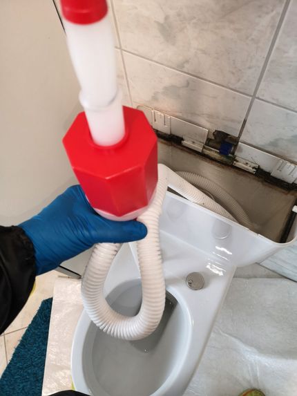 Person mit blauen Handschuhen benutzt einen rot-weißen Pömpel in einer weißen Toilette in einem Badezimmer. Der Pömpel ist in die Toilettenschüssel hineingeführt.