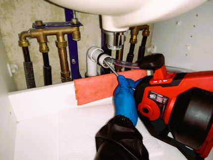 Eine Person mit blauen Handschuhen bedient eine Rohrreinigungsmaschine unter einem Waschbecken. Die Szene zeigt eine Sanitäranlage mit Rohren und Armaturen.