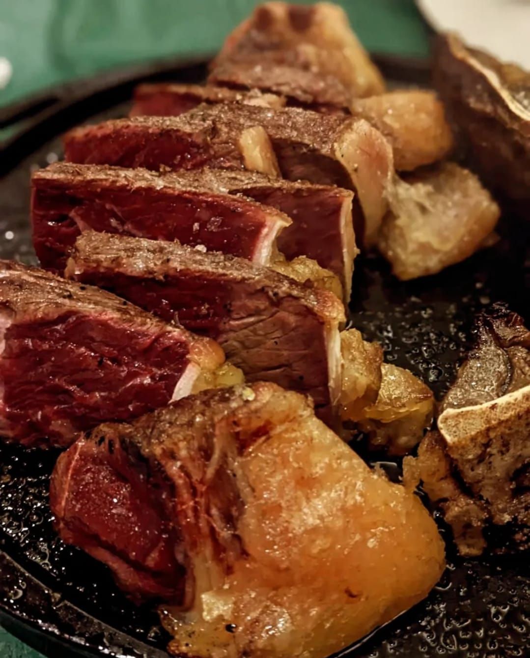Filete cortado en rodajas y sellado con trozos con hueso y patatas asadas en un plato negro.