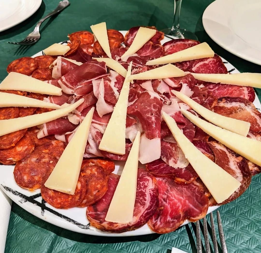 Un plato de chorizo en rodajas, jamón y cuñas de queso sobre una mesa con un mantel verde.