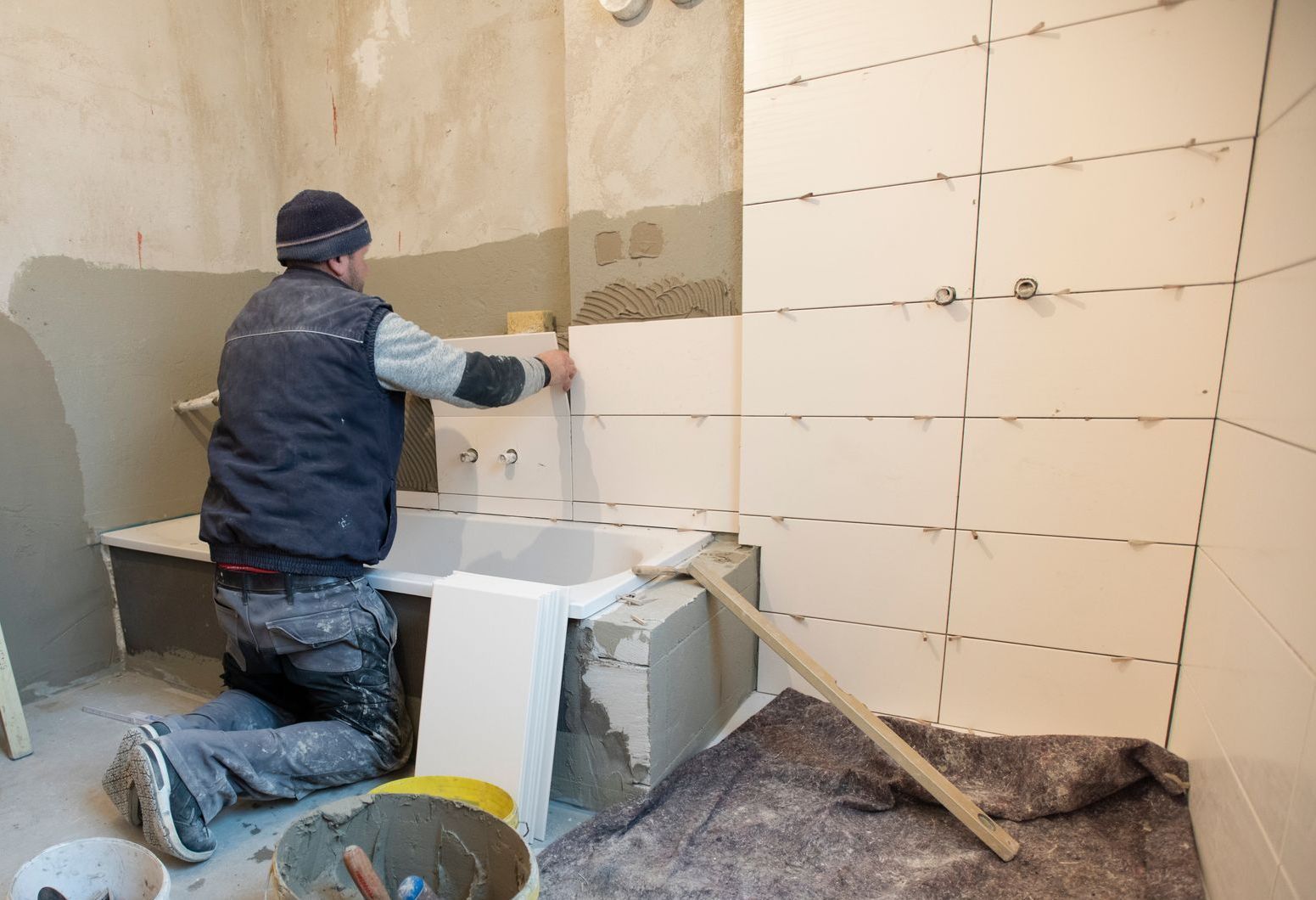 Mann fliest eine Badezimmerwand, kniet und legt eine Fliese. Helle Fliesen, grauer Zement und ein gelber Eimer.