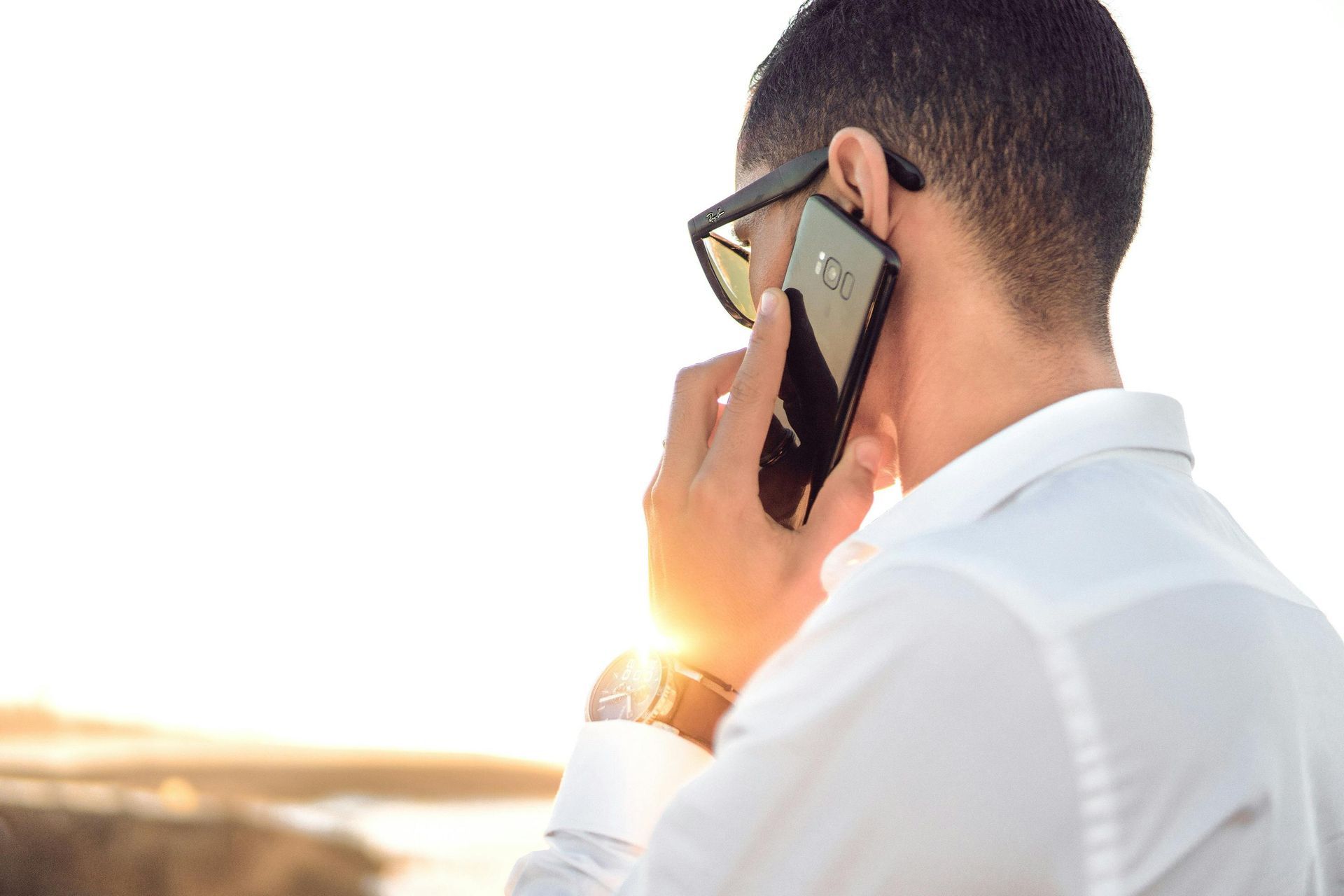 Mann in weißem Hemd, Sonnenbrille, hält ein Smartphone ans Ohr, draußen im Sonnenlicht.