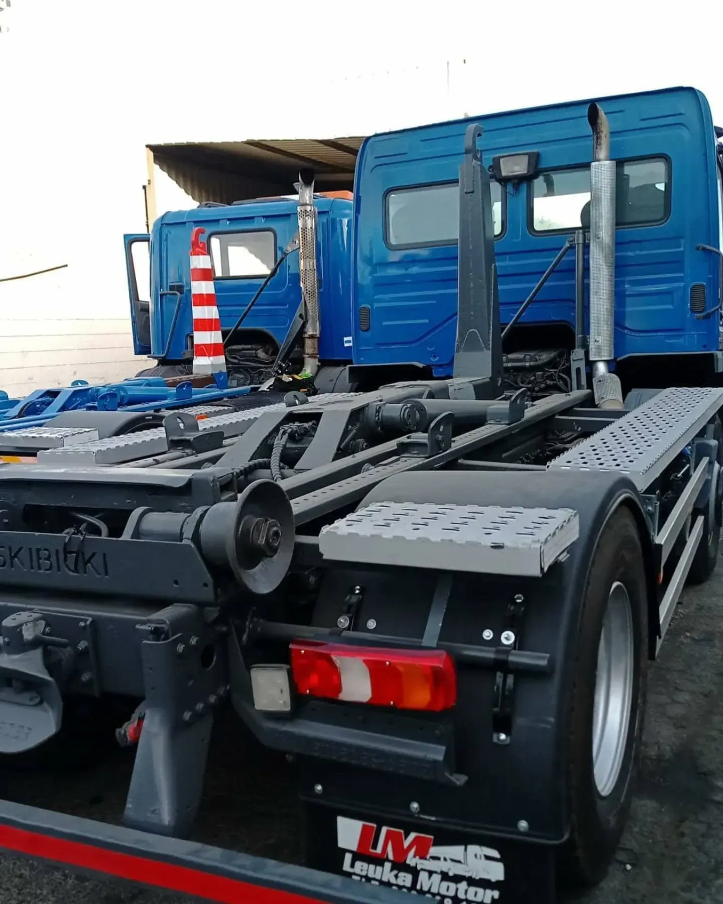 Camión con gancho elevador azul estacionado al aire libre, con un segundo camión detrás.