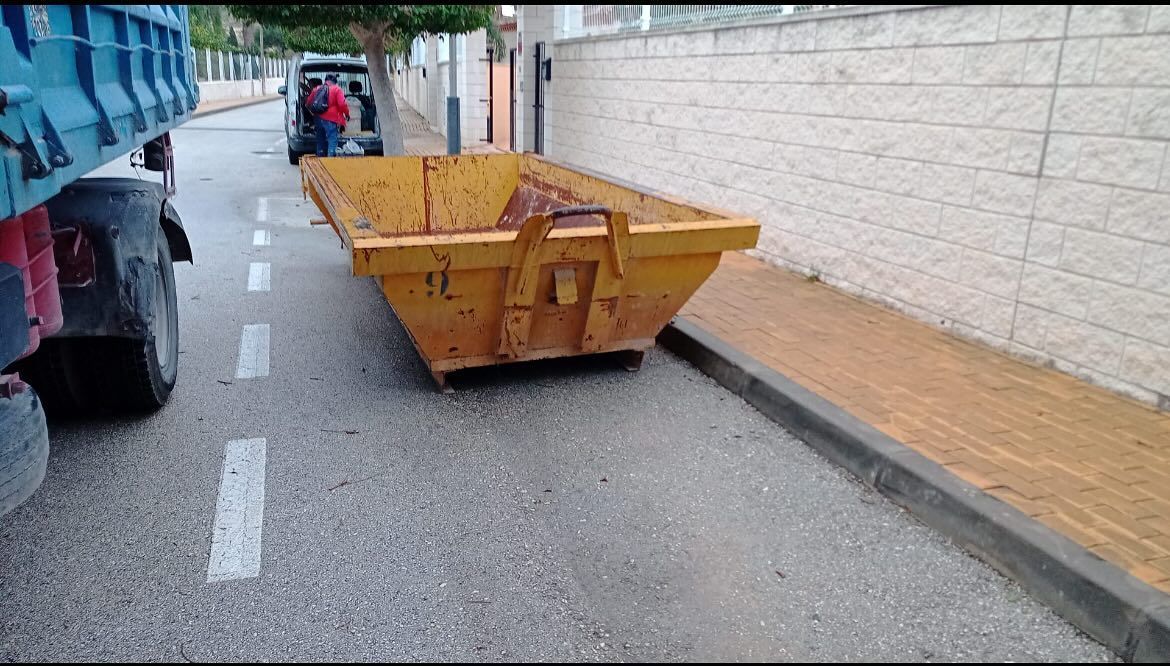 Contenedores verdes llenos de escombros de construcción en un área pavimentada, árboles y cielo de fondo.