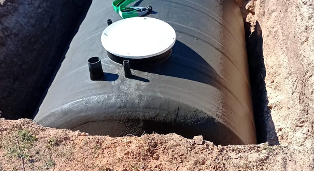 Obra con excavadoras cavando zanjas. Un trabajador con camisa amarilla y casco blanco trabaja cerca de un pozo lleno de agua.