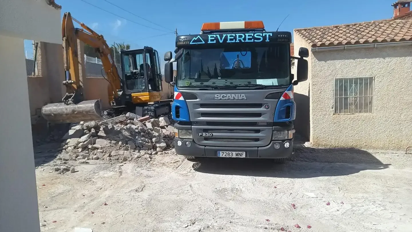 Una excavadora derriba el muro de un edificio, con escombros esparcidos debajo. Máquina amarilla, pared beige, cielo azul.