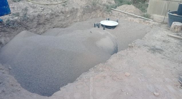 Camión volquete descargando una pila de grava azul sobre una superficie pavimentada cerca de árboles y tuberías.