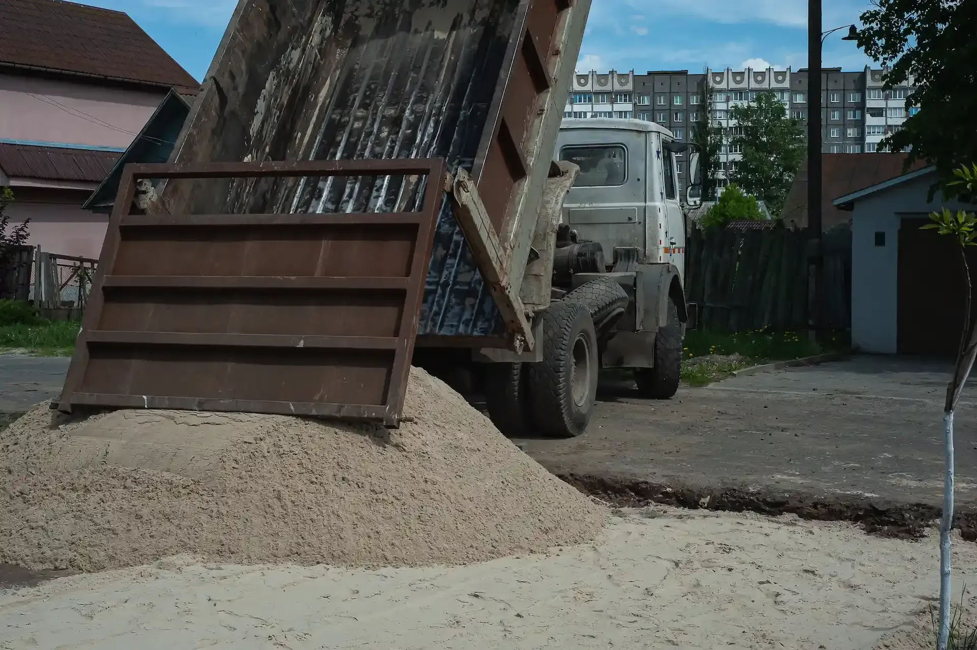 Camión volquete descargando grava en un camino de tierra, zona residencial al fondo.
