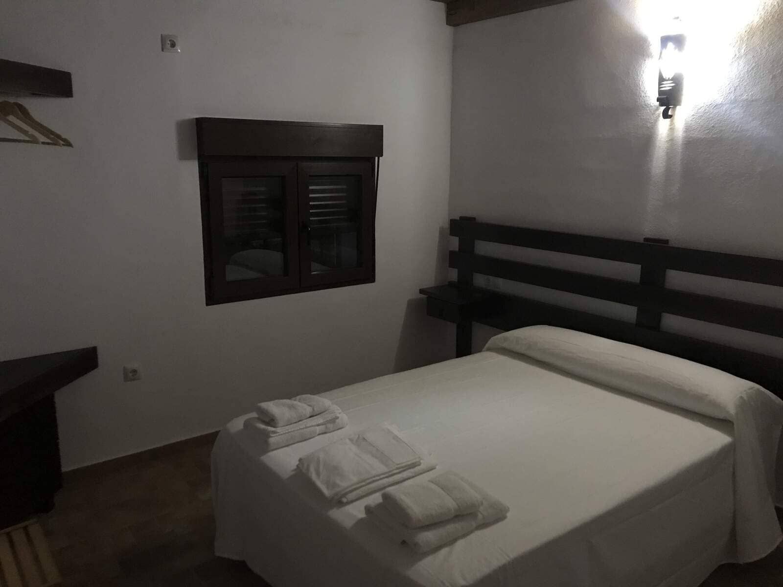 Dormitorio con ropa de cama blanca, toallas, cabecero de madera oscura, pequeña ventana y aplique de pared.