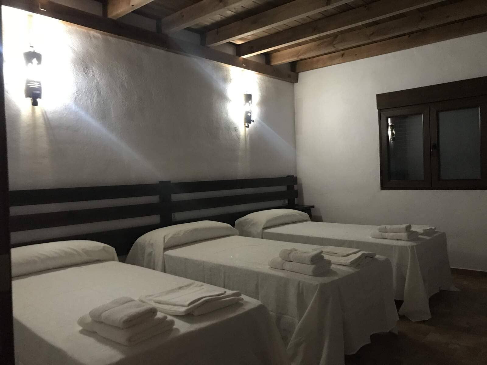 Dormitorio con tres camas individuales, sábanas blancas, cabecero de madera rústica, iluminación de aplique y una pequeña ventana.