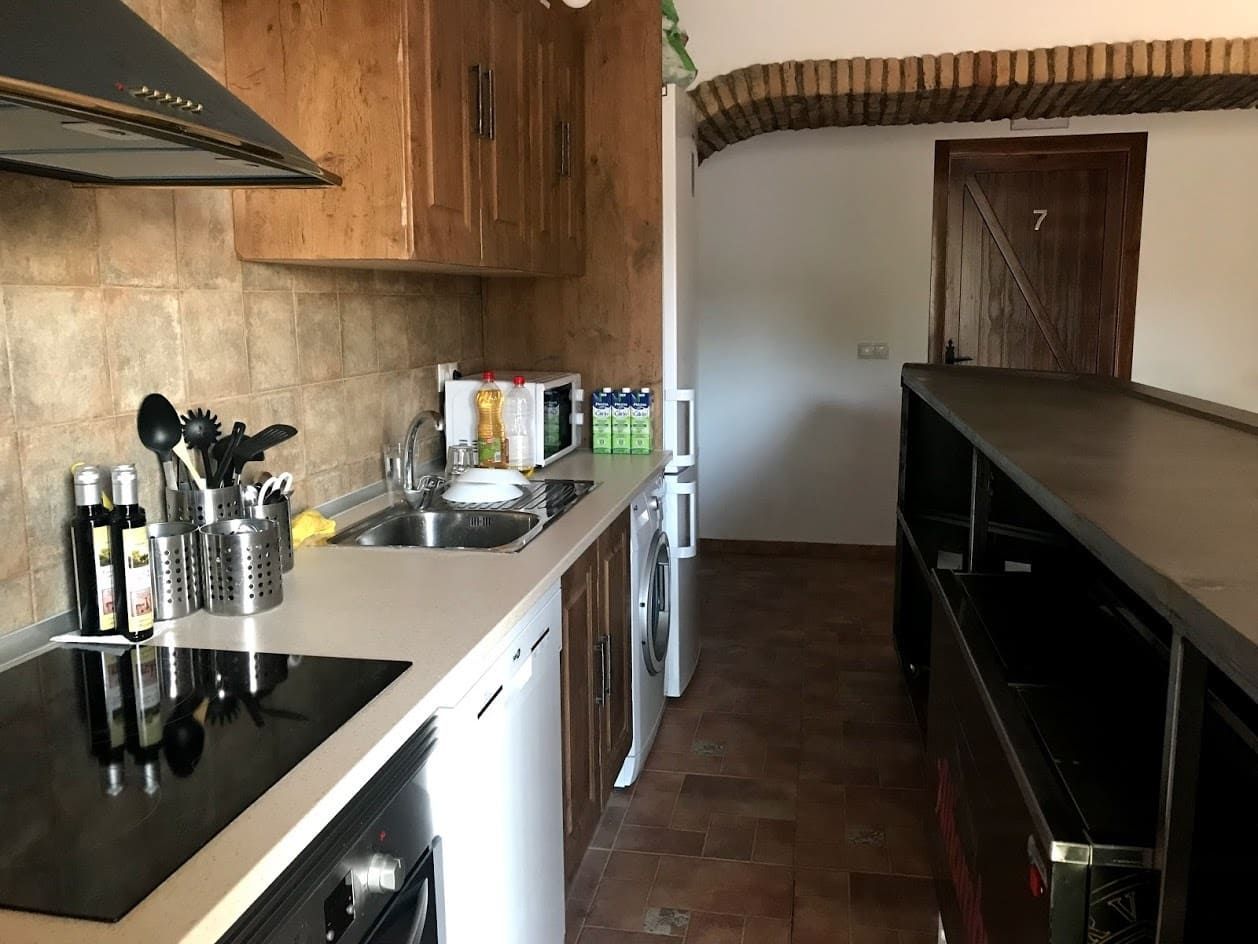 Cocina con gabinetes de madera, protector contra salpicaduras de color beige, electrodomésticos blancos y una encimera larga.