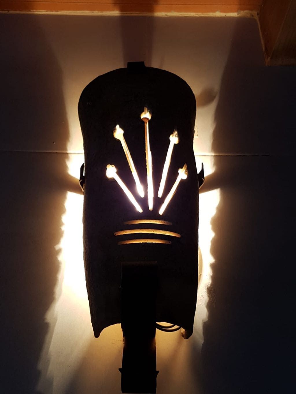 Aplique de pared de metal negro, que emite luz a través de flechas recortadas y un diseño de líneas horizontales, proyectando sombras.