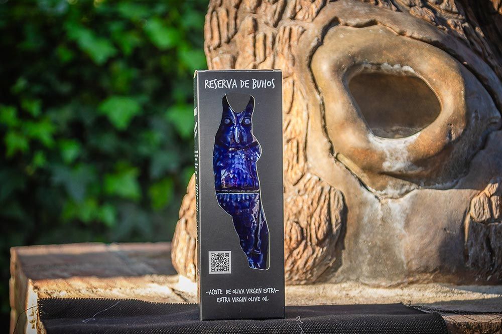 Una caja alta y oscura, con una escultura de cristal azul en forma de conejo, se alza sobre una superficie de madera frente a un tronco de árbol texturizado.