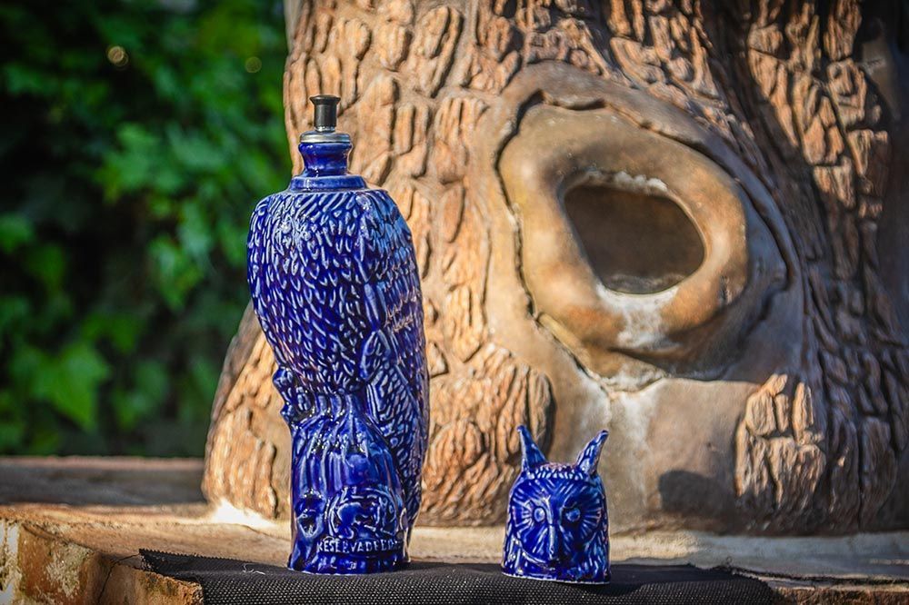 Una botella decorativa alta de color azul y una pequeña figurita de animal azul se encuentran al aire libre, frente a una estructura texturizada con forma de árbol.