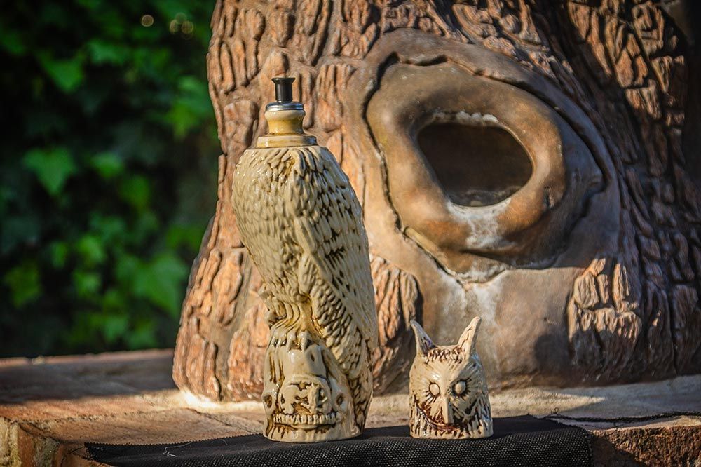 Dos adornos tallados con forma de búho, uno alto y otro pequeño, reposan sobre una superficie de madera frente a una escultura tallada en un tocón de árbol.