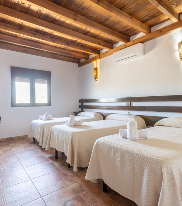Dormitorio con tres camas individuales, sábanas blancas, cabeceros de madera y vigas del techo a la vista.