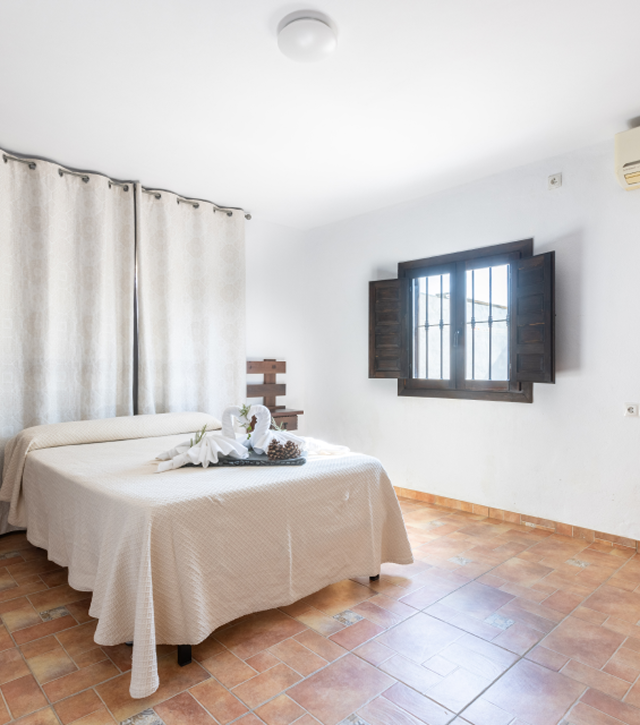 Dormitorio con cama doble, ventana abierta y toallas decorativas. Paredes y suelos en tonos claros.