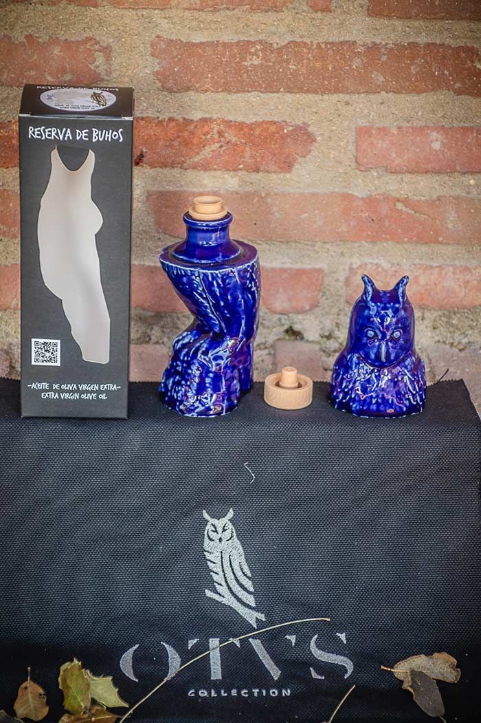 Una jarra azul con forma de búho y su tapón a juego, junto a su embalaje negro, se exhiben frente a una pared rústica de ladrillos.