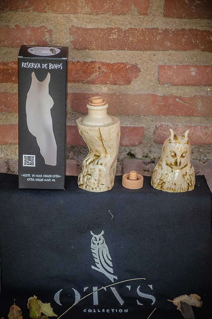 Una botella de cerámica con forma de búho, su cabezal/tapón extraíble y su caja negra con el logotipo de la marca, apoyada contra una pared de ladrillos.